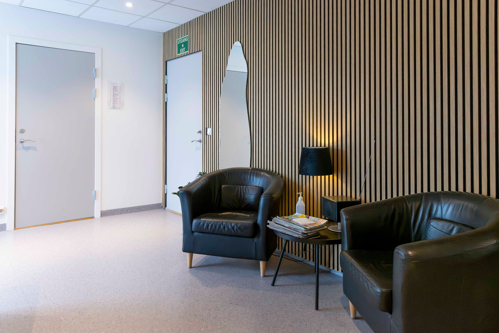 Sittegruppe i venterommet med to svarte skinnlenestoler, lite bord med magasiner og gulvlampe foran trespileveggen