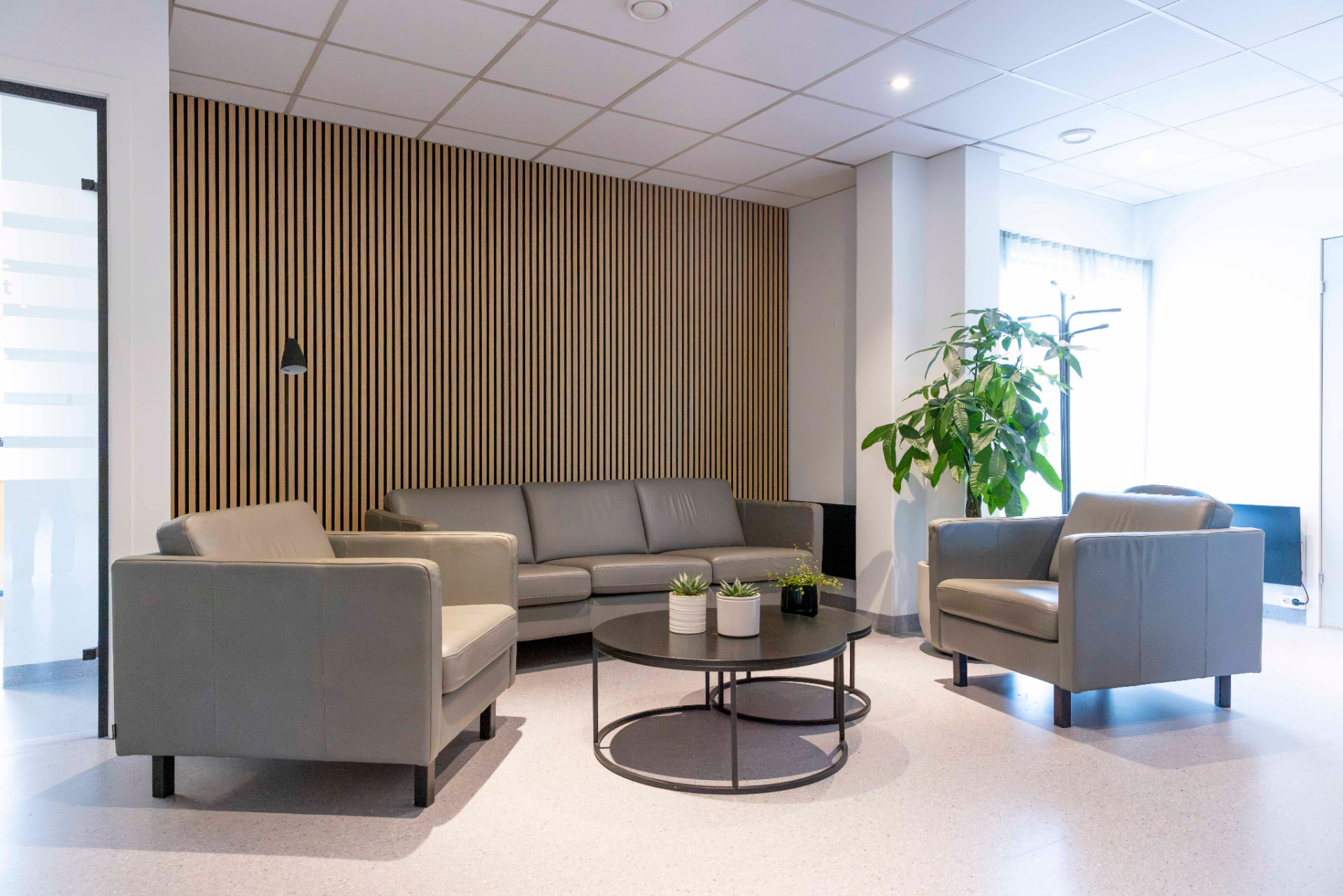 Venterommet med grå sofa og lenestoler rundt et rundt sofabord, trespilevegg og stor grønn plante ved vinduet